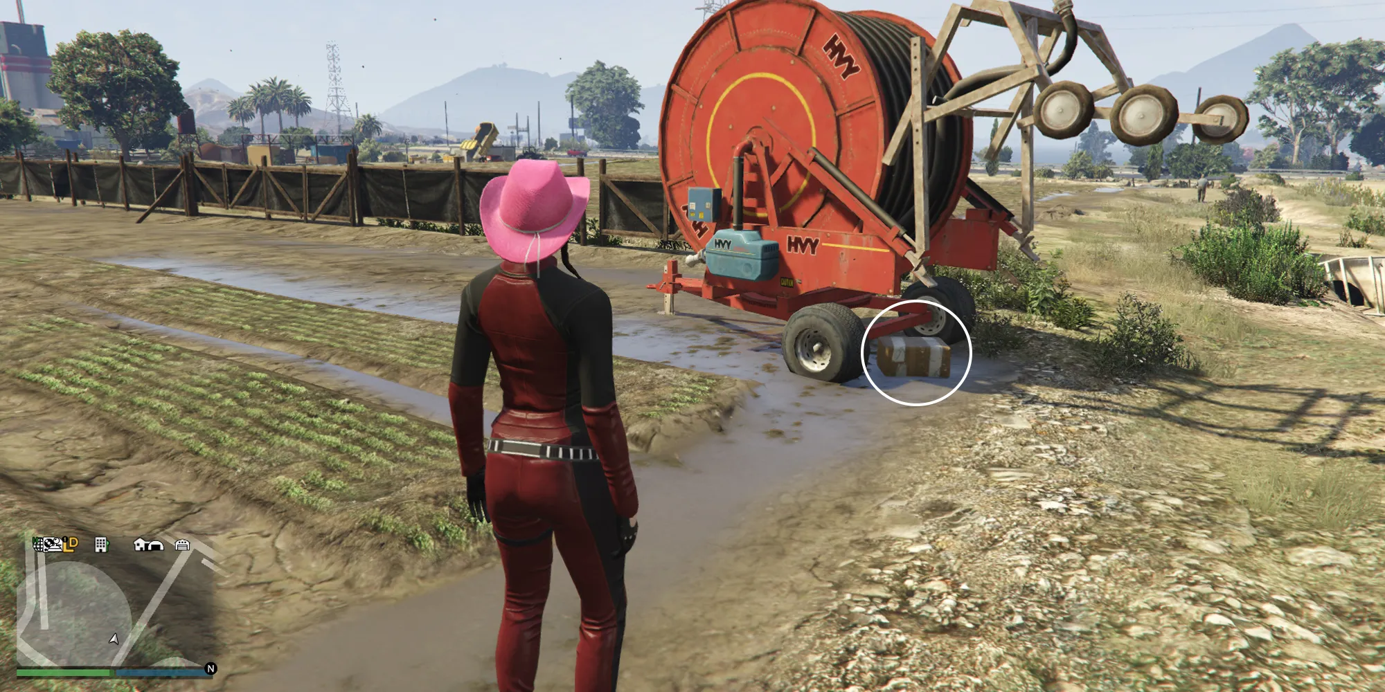 GTA OnlineのGrapeseed Rail Terminusにいる女性キャラクターが、農場のフィールドにある赤い農業機械の前に立っています。機械の前のタイヤの下にはGのキャッシュボックスがあります。