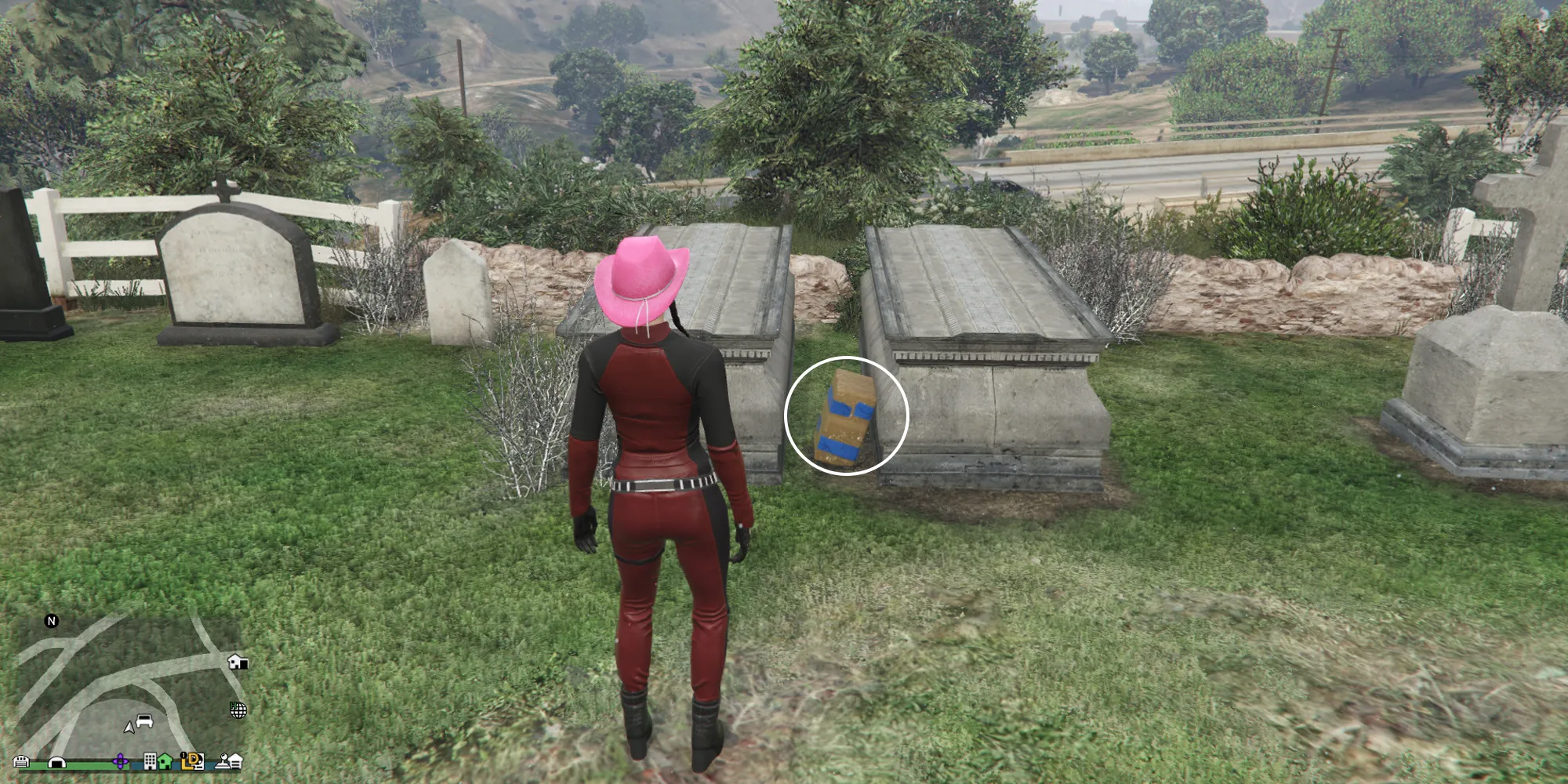 GTA Onlineの女性キャラクターが、教会の墓地にある2つの墓の前に立っています。周りには他の墓石や記念碑があります。大きな墓の間にGのキャッシュがあります。