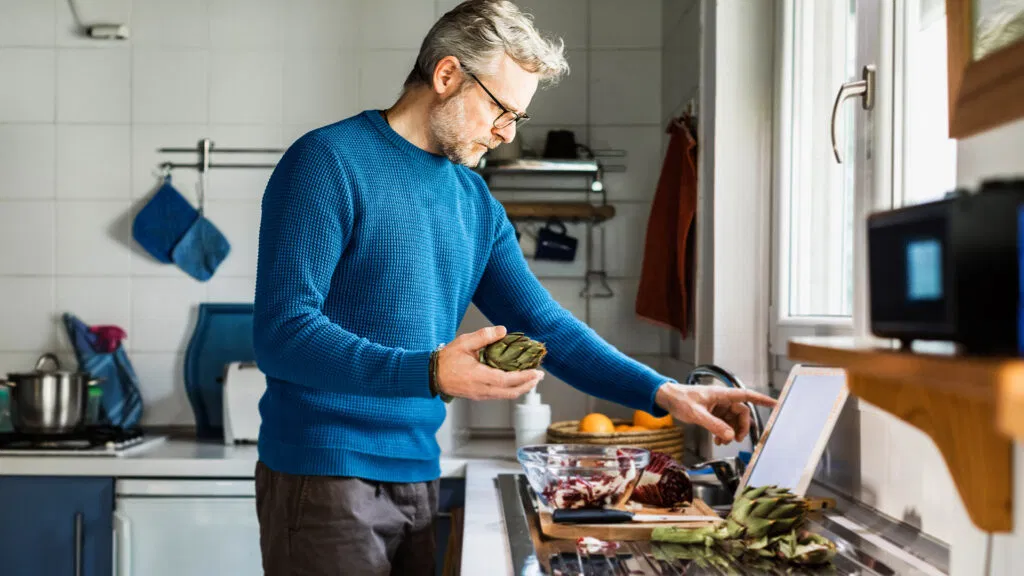 Un homme tient un artichaut dans sa cuisine en lisant une recette