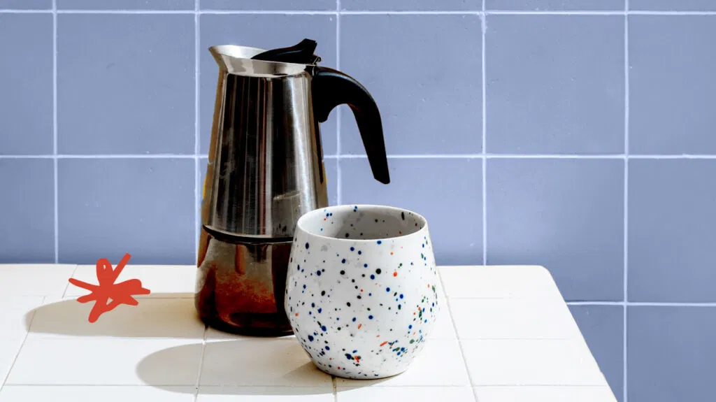 metal coffee pitcher and white cup with colorful speckles against a blue tile background