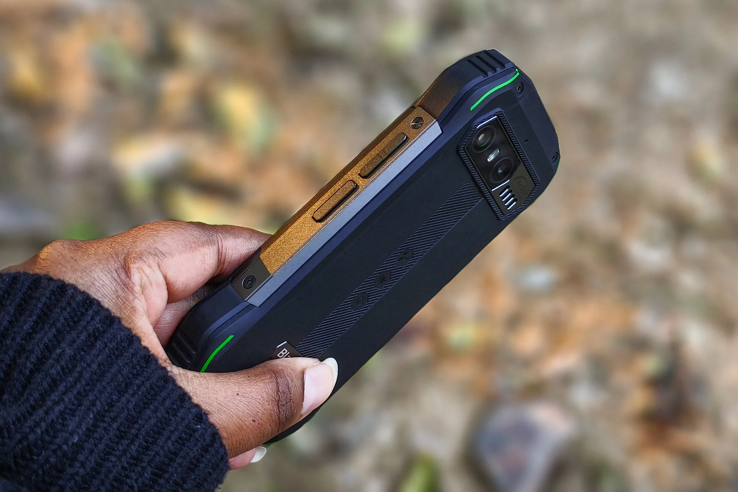 Persona sosteniendo el pequeño teléfono Android resistente Blacview N6000 en la mano contra hojas secas en el suelo.