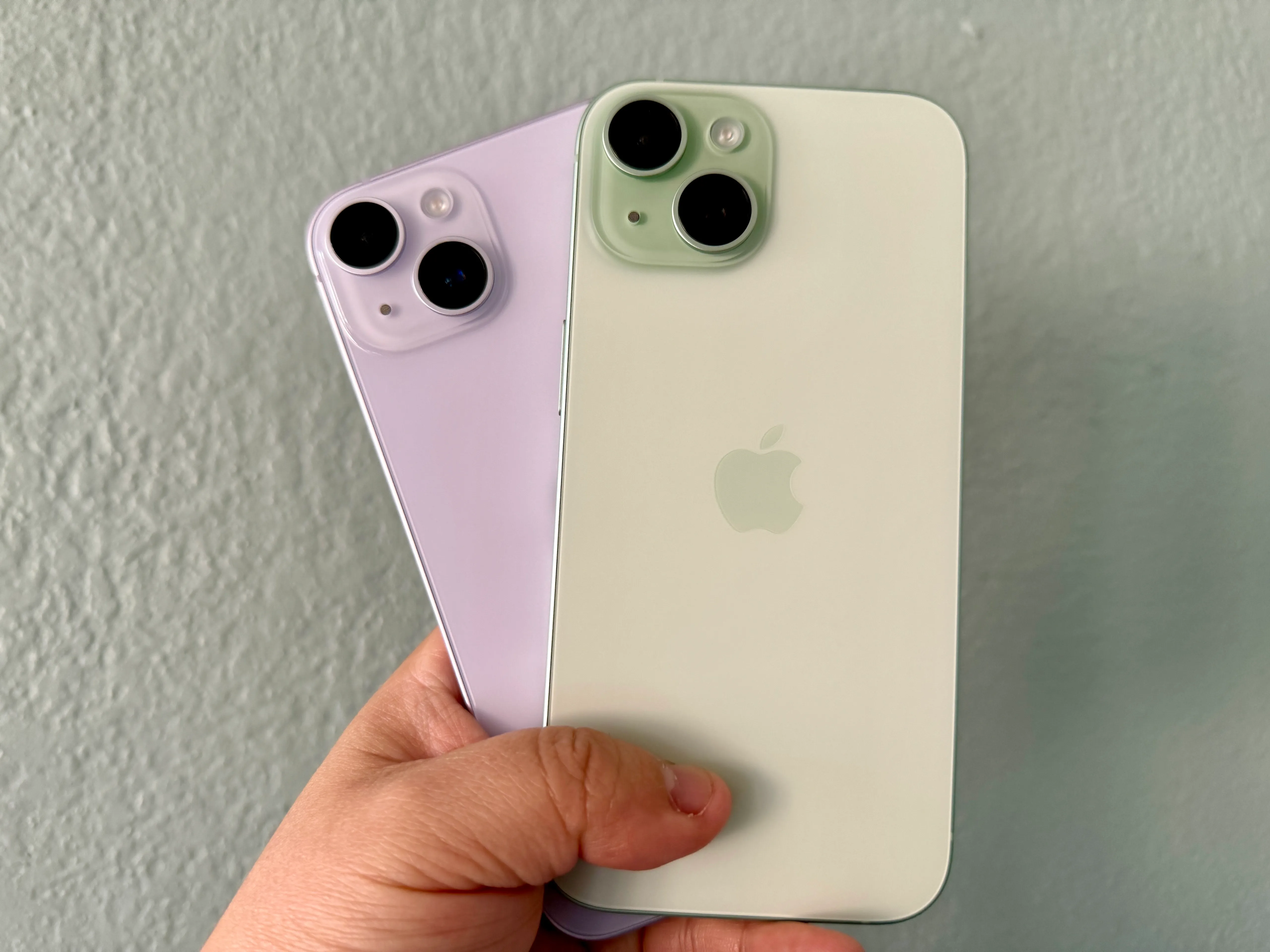 紫色的iPhone 14（左）和手里拿着一个绿色的iPhone 15。