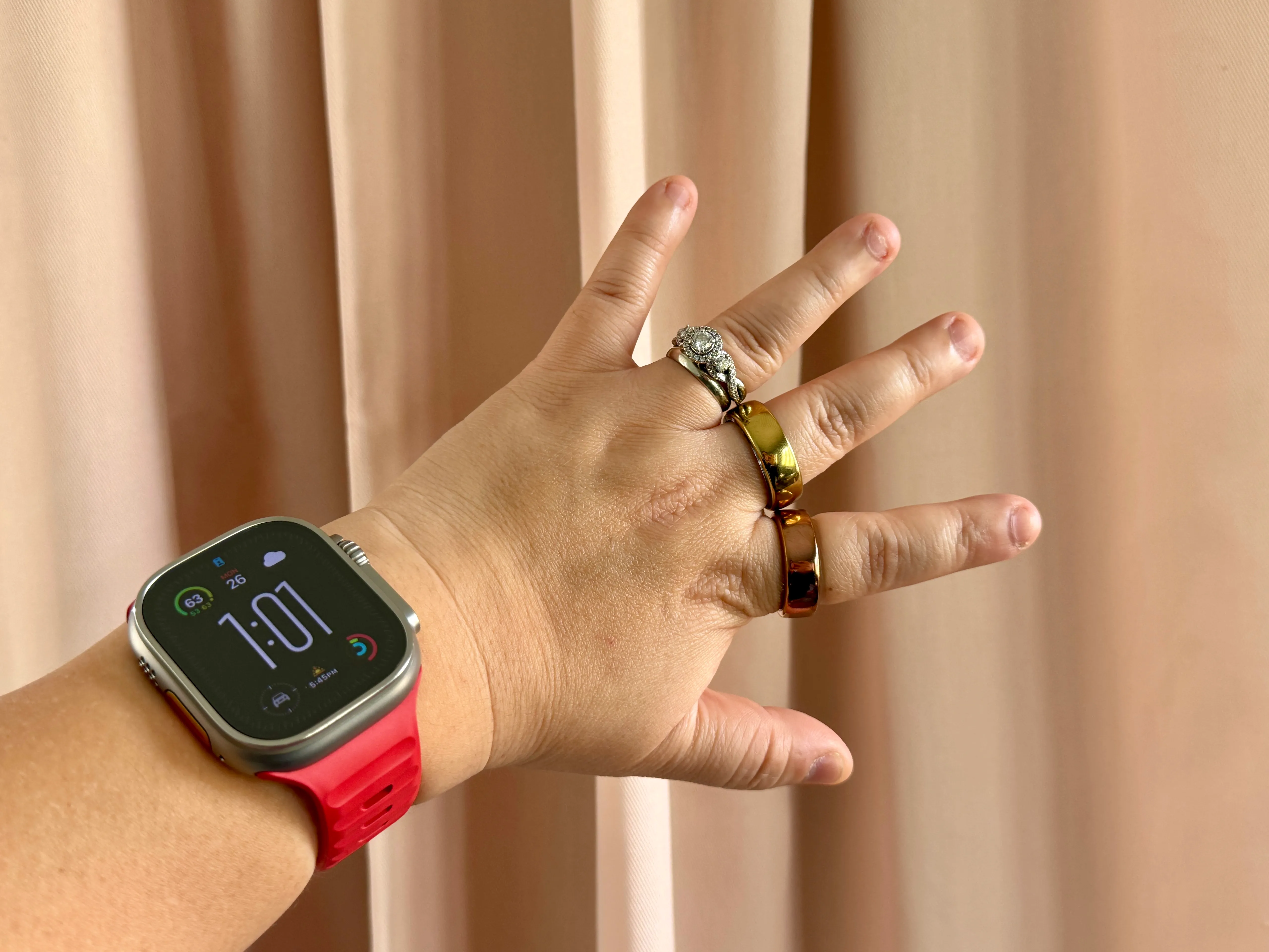 戴有红色表带的 Apple Watch Ultra，手指上有 Movano Evie 和 Oura 戒指。